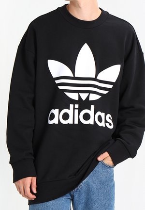 Personne portant un sweat-shirt noir avec un grand logo trèfle Adidas blanc et un texte, associée à un jean bleu, main dans la poche.