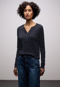 Vrouw met krullend donker haar draagt een marineblauw Henley shirt met lange mouwen en een blauwe spijkerbroek, staand tegen een effen lichtgrijze achtergrond.