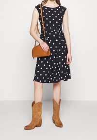 Kvinna iklädd en svart knälång klänning med polka prickar, bär på en brun axelväska och har på sig ljusbruna suède cowboystövlar.