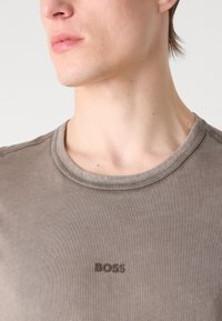 Szürke pamut póló, lekerekített nyakkivágással, a mellkasán egy diszkrét fekete "BOSS" logóval, sima, puha textúrával.