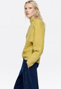 Musztardowy, puszysty sweter z wysokim dekoltem, w zestawie z ciemnoniebieskimi spodniami o szerokich nogawkach. Tekstura wydaje się miękka i przytulna.