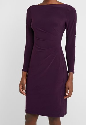 Strakke paarse jurk tot op de knie met lange mouwen, voorzien van zilveren knopen van de schouder tot de manchetten, gedragen door een staand persoon.