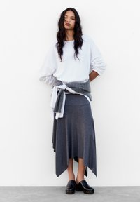 Weißer, übergroßer Pullover, kombiniert mit einem grauen Sweater um die Taille, dazu ein grauer asymmetrischer Rock und schwarze Holzclogs. Glatte Texturen, lässiges Design.