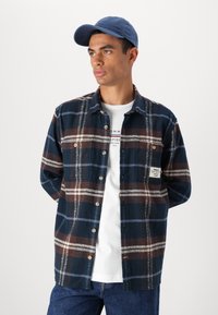 Camisa de flanela xadrez em azul escuro, castanho e branco; apresenta três botões à frente e um patch com o logo no bolso esquerdo. Usada sobre uma camisa branca.