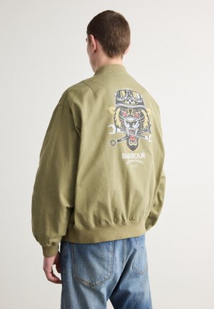 Persoon draagt een olijfgroene jas met op de achterkant een afbeelding van een brullende tijgerkop en gekruiste steeksleutels, gecombineerd met een blauwe spijkerbroek.