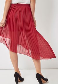 Falda midi plisada roja con un diseño fluido y una textura transparente, acabada con una cinturilla de satén y combinada con zapatos de tacón altos negros.