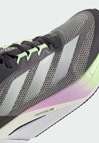 Baskets sportives avec une tige en mesh noire et grise, agrémentée d'accents verts. Design à trois bandes emblématique et semelle dégradée allant du violet au blanc.