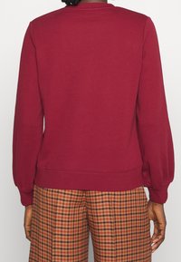 Sudadera burdeos con cuello redondo, puños acanalados y sutiles mangas abullonadas. Combinada con pantalones de cuadros en tonos marrón y naranja.