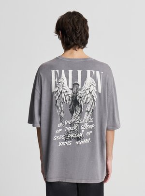 Persona che indossa una maglietta grigia oversize con ali bianche e la scritta "FALLEN" e una citazione sul retro riguardante gli dèi che sognano di essere umani.