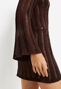 Καφέ πλεκτό φόρεμα με κάθετες διαφανείς ρίγες, με μακριά μανίκια και άνετη εφαρμογή. Η υφή του υφάσματος προσθέτει βάθος στο σχέδιο.