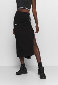 Černá sportovní sukně s vysokým rozparkem, s bílým logem Kappa a kontrastním bočním vzorem. Kombinováno s odpovídajícím crop topem a teniskami.