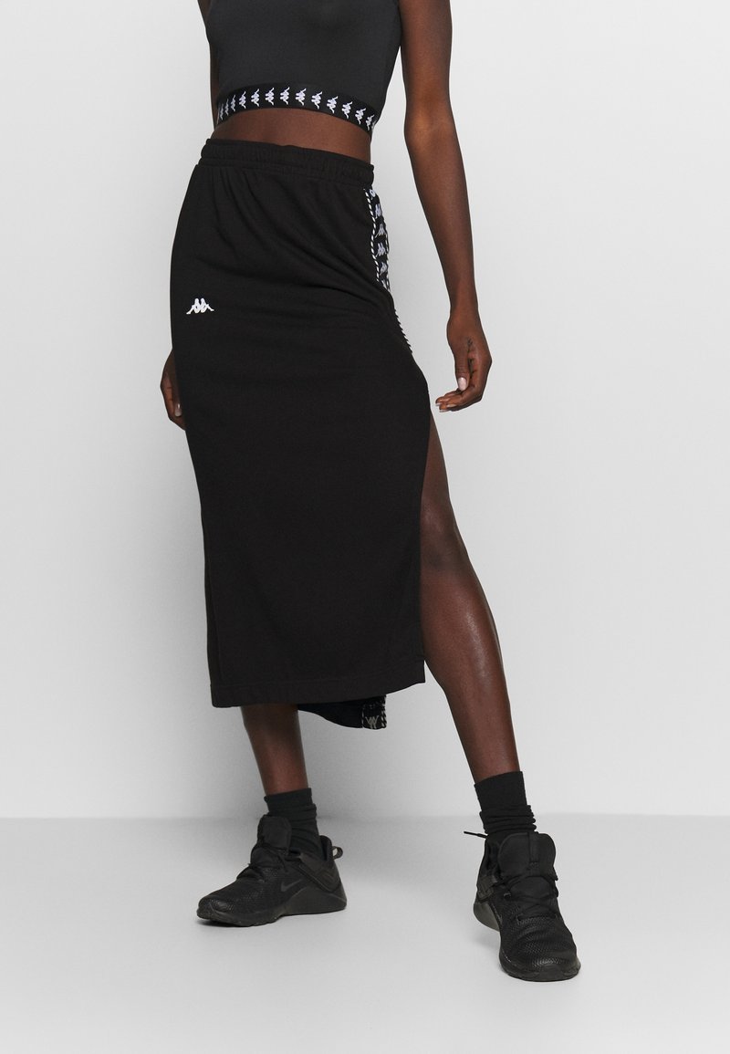 Černá sportovní sukně s vysokým rozparkem, s bílým logem Kappa a kontrastním bočním vzorem. Kombinováno s odpovídajícím crop topem a teniskami.