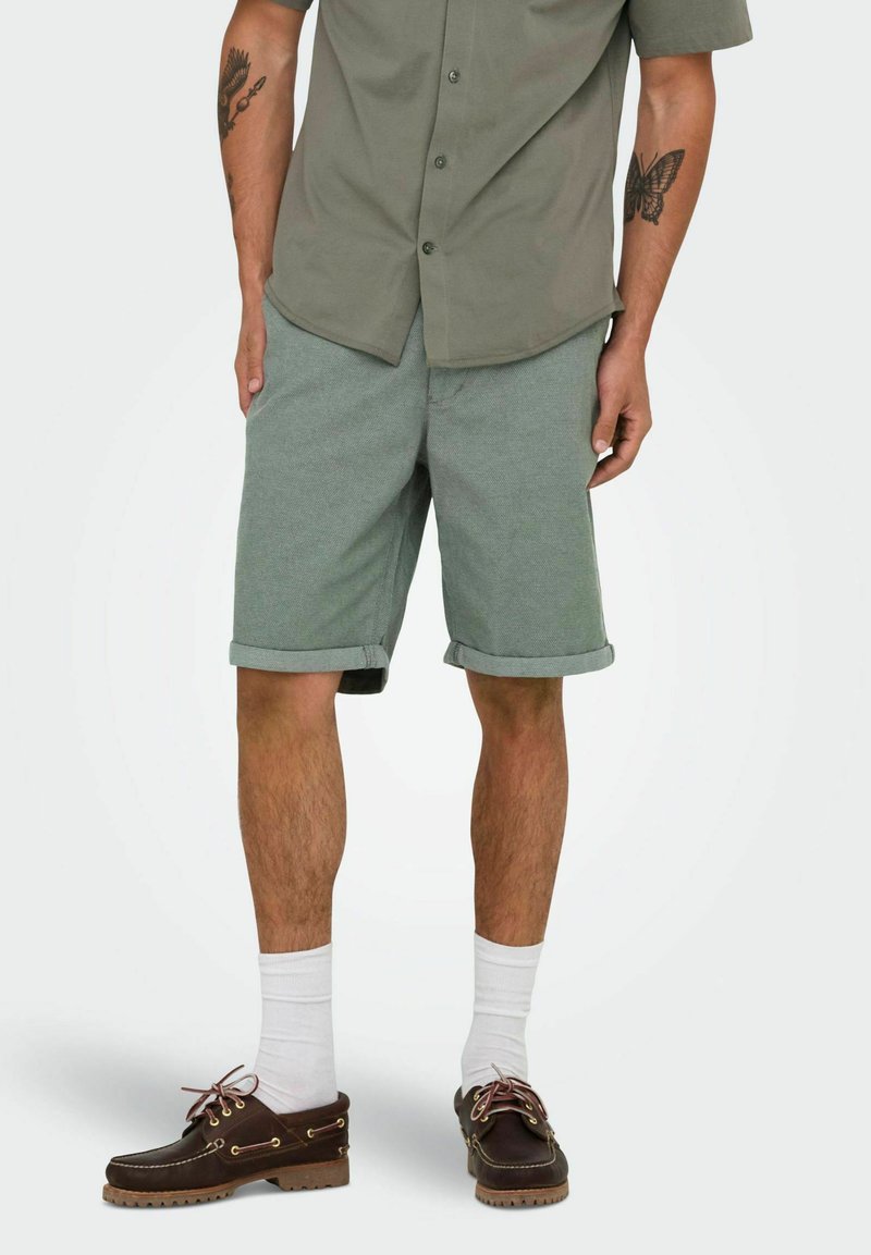 Grüne Baumwollshorts mit aufgekrempelten Säumen, kombiniert mit einem grauen Hemd und braunen Schuhen. Enthält weiße Socken und ein Schmetterlingstattoo.
