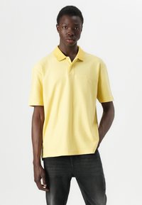 Gul polo-skjorte med strukturert krage og korte ermer, med en subtil logo på brystet og en avslappet passform. Brukt sammen med mørke jeans.