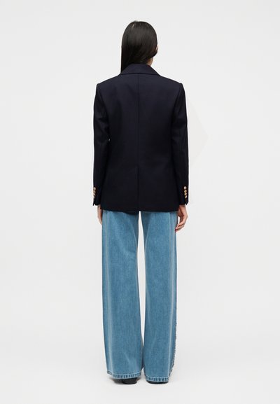 Γυναίκα με μακριά σκούρα μαλλιά, ντυμένη με μπλε σκούρο σακάκι με χρυσά κουμπιά και φαρδιά ανοιχτό μπλε τζιν, στέκεται αντικρίζοντας έναν απλό λευκό τοίχο.