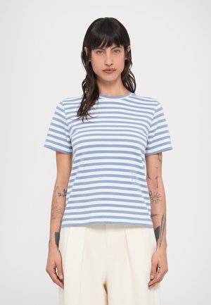 Άτομο με σκούρα μαλλιά και τατουάζ που φορά μπλε και λευκό ριγέ μπλουζάκι και λευκό παντελόνι σε απλό φόντο.