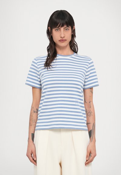 Personne aux cheveux foncés et tatouages portant un t-shirt rayé bleu et blanc et un pantalon blanc sur un fond uni.