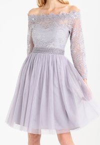 Šaty v levandulové barvě s odhalenými rameny, krajkovým korpusem, průhlednými dlouhými rukávy a plisovanou tylovou sukní. Beadovaný pás přidává detail.