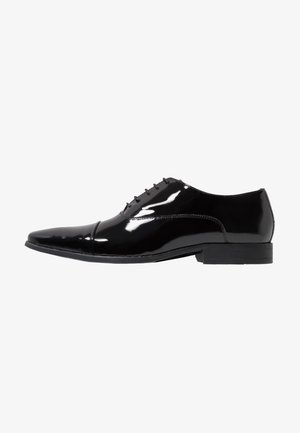 Chaussure habillée en cuir verni noir avec un bout pointu, six œillets et une semelle en caoutchouc plate. Fini lisse et brillant avec des détails de couture minimalistes.