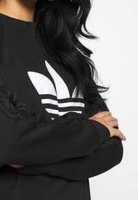 Svart sweatshirt med en stor vit Adidas-logotyp, dekorativ spetskant på ärmarna och ribbade manschetter, med en avslappnad passform.