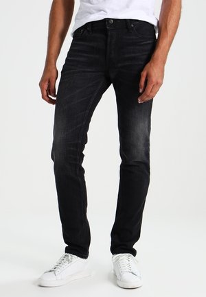 Muž stojící před bílým pozadím, oblečený v úzkých černých džínách a bílých teniskách, ruce volně podél těla.