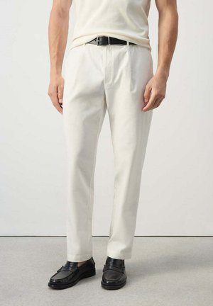 Hombre con pantalones blancos, cinturón negro y mocasines de cuero negro, de pie frente a un fondo claro y liso.