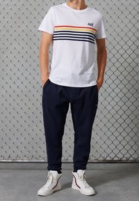 Vit bomullströja med flerfärgade horisontella ränder; parat med marinblå joggerbyxor och vita högskurna sneakers med röda detaljer.