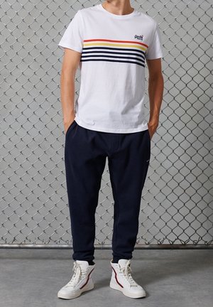 Camiseta blanca de algodón con rayas horizontales multicolores; combinada con pantalones jogging azul marino y zapatillas altas blancas con acentos rojos.