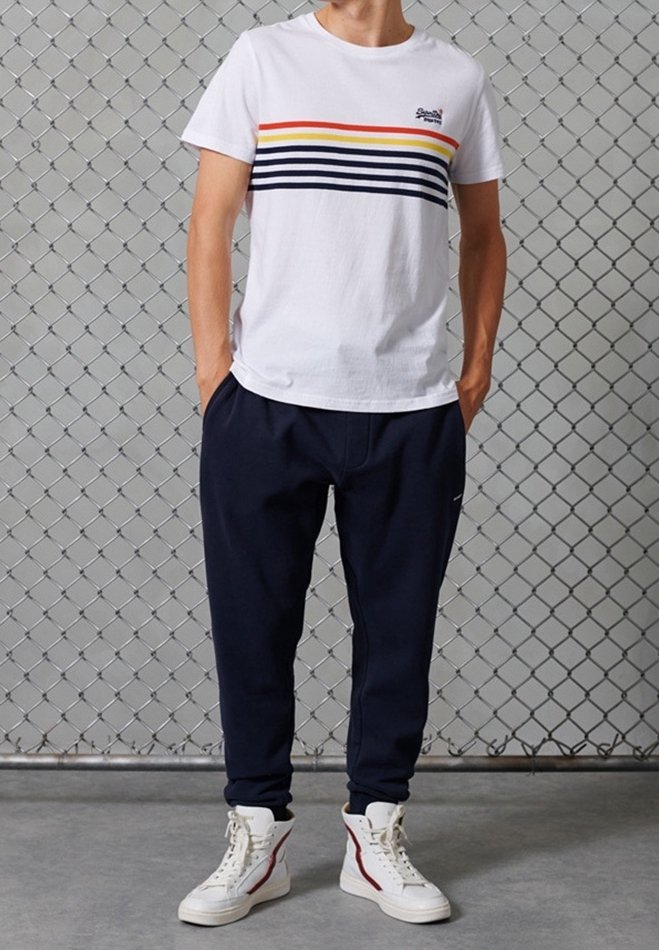 Vit bomullströja med flerfärgade horisontella ränder; parat med marinblå joggerbyxor och vita högskurna sneakers med röda detaljer.