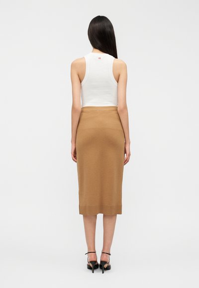 Λευκό ριμπ μπλουζάκι χωρίς μανίκια, πλεκτή midi φούστα σε ανοιχτό καφέ χρώμα, λεία υφή, εφαρμοστή σιλουέτα και λογότυπο στο πίσω μέρος. Μαύρα πέδιλα με τακούνι.