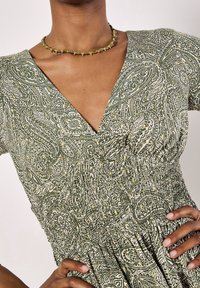 Green patterned dress with a deep V-neck and smocked waist. Small yellow accents throughout. Worn with a simple accessory necklace.