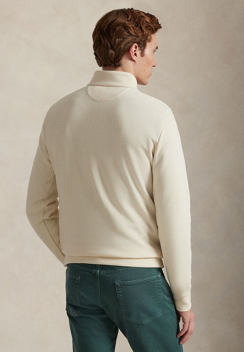 Young man seen from behind wearing a cream-colored long-sleeve pullover and dark green pants against a neutral background.