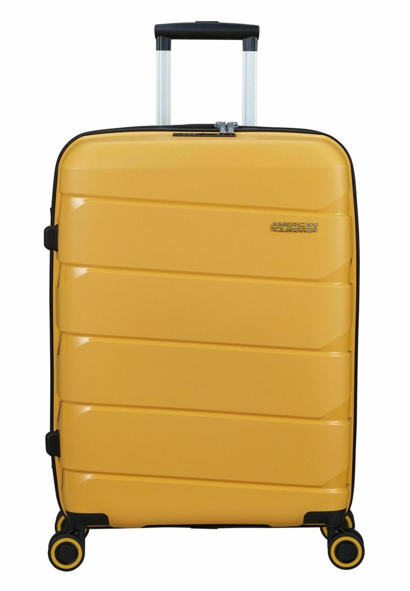 Harde gele koffer met horizontale ribbelstructuur, zwarte accenten, telescopische handgreep en vier zwarte wielen. Merklogo zichtbaar.