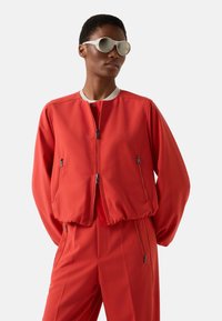 Rød bomberjakke med ribbet krave, lynlåslukning foran og sidelommer. Glat stof, afslappet pasform, kombineret med matchende røde bukser.