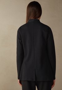 Czarna długa kurtka z długim rękawem, z wysokim kołnierzem, z szwem z tyłu i lekko rozszerzonym dołem. Gładka tekstura z minimalnymi detalami w designie.