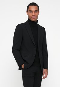 Črn moški suknjič s slim fit krojem in enojnim zapenjanjem. V kombinaciji s črnim turtleneckom, ki prikazuje gladko, teksturirano blago.
