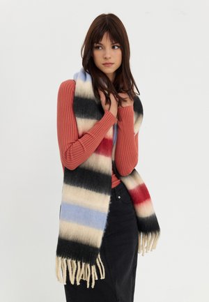 Striped scarf with blue, beige, red, and black, made of soft material, featuring fringed edges. Model wears it with a ribbed sweater.