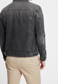 Chaqueta de mezclilla negra con un acabado desgastado, presenta un cuello, mangas largas y dos detalles de botones a presión en la cintura. Diseño minimalista.
