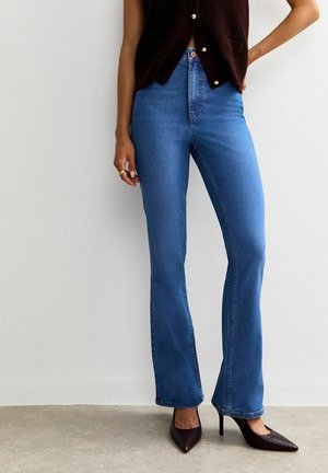 Jean bleu coupe slim porté avec des chaussures à talons noires et un haut sans manches foncé avec des boutons, debout contre un mur blanc.