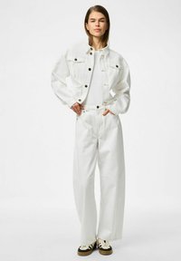 Vit denimjacka med svarta knappar, i kombination med vita högmidjade vida byxor. Modellen bär en åtsittande vit topp och avslappnade sneakers.