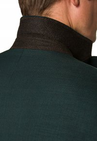 Zielona teksturowana kurtka z filcowym kołnierzem w ciemnym brązie. Widoczne detale szwów wzdłuż kołnierza i szwu na ramieniu.