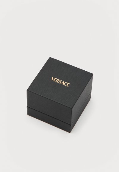 Boîte carrée texturée noire avec le logo Versace doré centré sur le couvercle, placée sur une surface blanche unie.