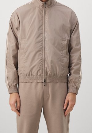 Person trägt taupefarbene Reißverschlussjacke mit elastischen Bündchen und passenden Hosen, steht vor hellem, einfarbigem Hintergrund.