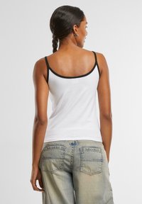 Hvit tanktopp med svart kant, laget av myk tekstil. Baksiden har rund hals og tynne stropper, kombinert med lyseblå jeans.