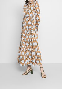 Robe à manches longues avec boutons, motif floral en orange et bleu, dotée d'une ceinture à la taille et d'une jupe à volants. Associée à des sandales vertes.