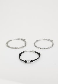 Trois bracelets : deux chaînes en argent avec des styles de maillons différents et un bracelet en cordon noir avec un détail de boucle en argent.