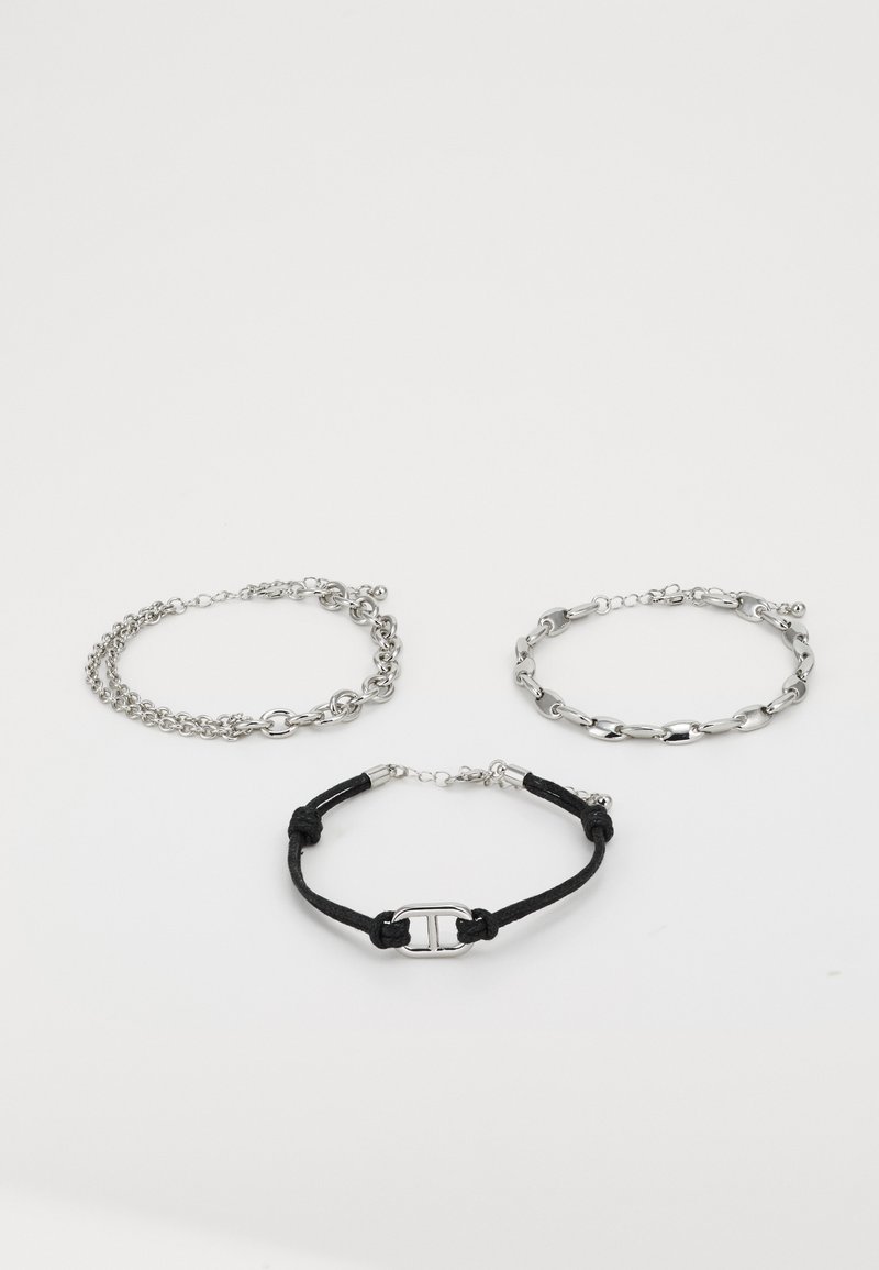 Trois bracelets : deux chaînes en argent avec des styles de maillons différents et un bracelet en cordon noir avec un détail de boucle en argent.