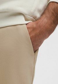 Sima textúrájú bézs nadrág, oldalsó zsebbel, amelyben egy kéz pihen, világos színű bordázott pulóverrel kombinálva.