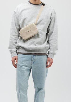 Béžová taška přes rameno s texturovaným předním dílem, zapínáním na zip a nastavitelným popruhem, doplněná o šedou mikinu a světle modré džíny.