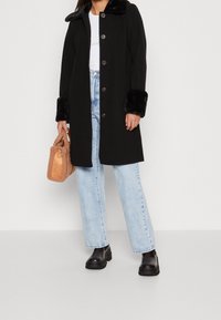 Manteau noir avec col et poignets en fausse fourrure, six boutons, porté sur un haut blanc et un jean bleu clair, associé à des chaussures à plateforme noires.